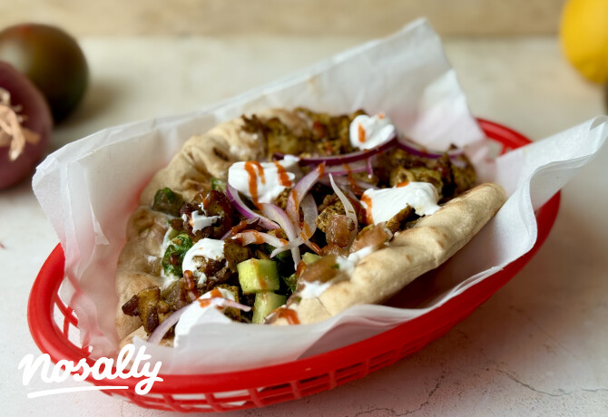 Pita kenyérbe töltött vegán gyros zöldségekkel és pirított növényi falatokkal, tetején krémes fehér szósszal és csípős szósszal meglocsolva. A szendvics papírral bélelt piros kosárban van tálalva, friss uborkával és lilahagymával.