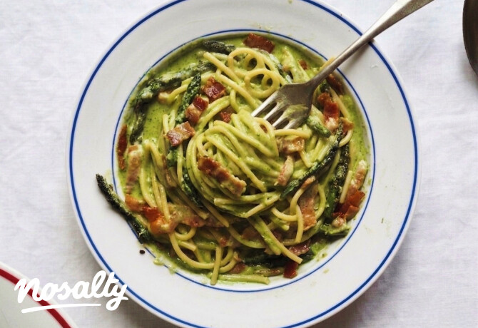 Tányérban tálalt spárgás carbonara felülnézetből: krémes, zöld spárgaszósszal bevont spagetti ropogósra sült szalonnadarabokkal és pirított spárgafejekkel, villával a tészta közepén.