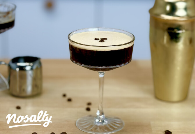 Espresso Rumtini koktél elegáns kehelypohárban, világos krémhabbal és három kávészemmel díszítve. A háttérben aranyszínű koktélshaker, kávékiöntő és elszórt kávészemek láthatók egy fa munkalapon.