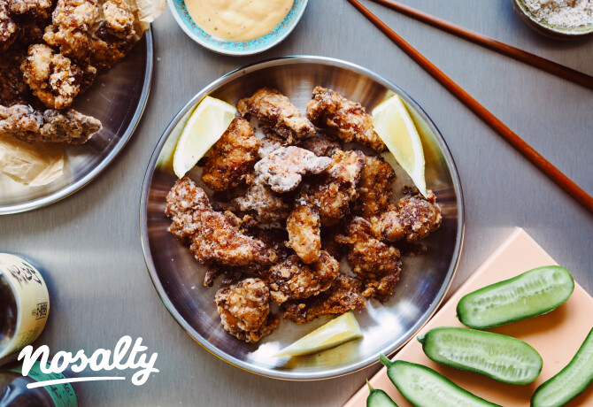 Fém tányéron tálalt ropogós japán karaage csirkefalatok citromgerezdekkel, mellette uborkaszeletek, pálcikák és egy tál krémes mártogatós egy világos asztalon.
