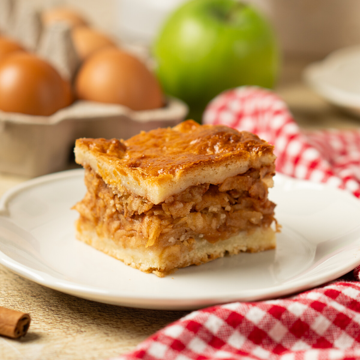 Almás pite kockára vágva, fehér kistányéron, mellette kockás konyharuha