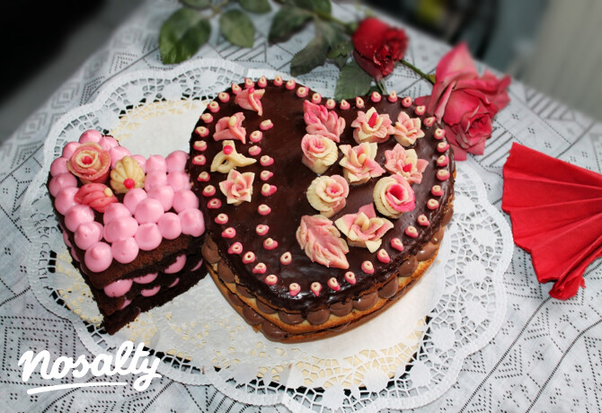 Kerek, csokoládémázas torta fondant rózsákkal, apró cukordíszekkel, mellette egy szelet rózsaszín virágokkal díszített torta. A sütemények fehér csipketerítőn állnak, mögöttük vörös rózsák.