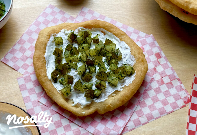 Kovászos uborkával és tejföllel megkent lángos rózsaszín-fehér kockás papíron, fa asztalon tálalva.