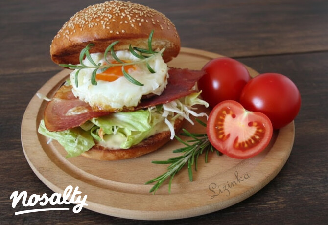 Szezámmagos hamburgerzsemlében tálalt reggelizőburger tükörtojással, ropogós baconnel, salátával és friss rozmaringgal, mellette egész és félbevágott paradicsom egy fa tálcán