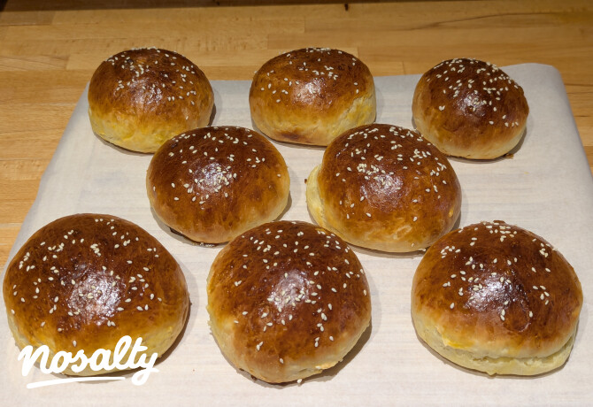 Kilenc aranybarnára sült, kerek burgonyás hamburgerbuci sorakozik egy fa deszkán, a tetejükön szezámmaggal.