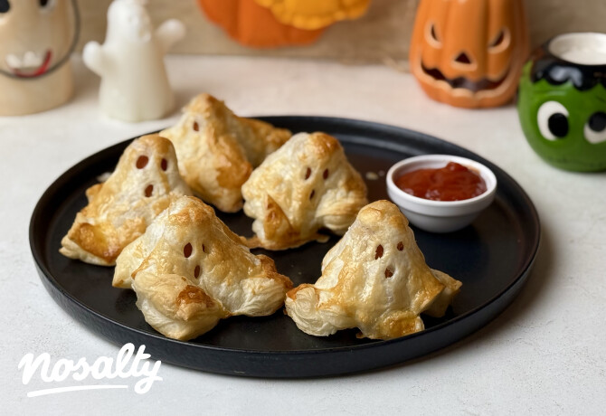 Aranybarnára sült, virslivel töltött leveles tésztás halloweeni szellemek kivágott szemekkel és szájjal, fekete tányéron, ketchup és tökös dekorációk mellett.