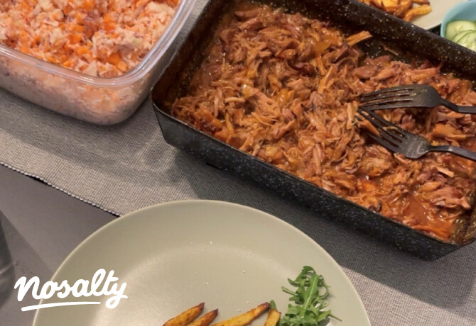 Tepsiben sült BBQ tépett hús látható villákkal, mellette egy dobozban friss káposztasaláta. Az asztalon tányéron hasábburgonya és egy kevés rukkola, a háttérben ketchupos szósz is látszik.